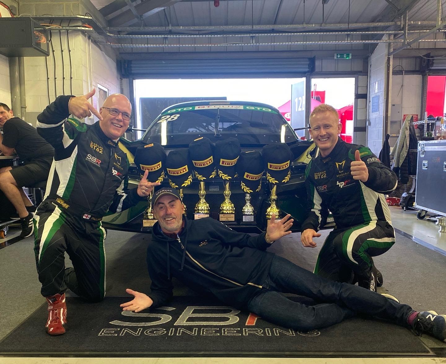 Three members of the SBR Engineering team in race suits celebrating with a row of trophies in front of the Brabham BT62