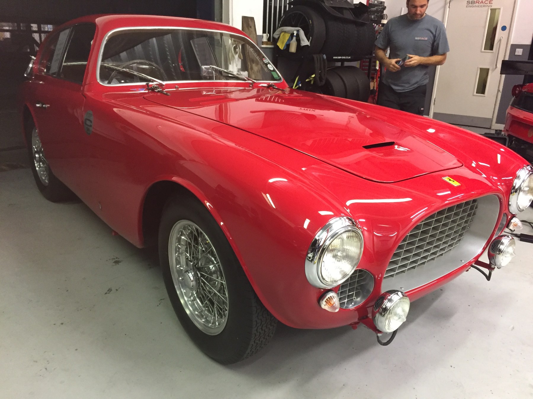 A red 1950s Ferrari 250 style coupe with wire wheels and chrome grille in the SBR workshop, with a technician behind