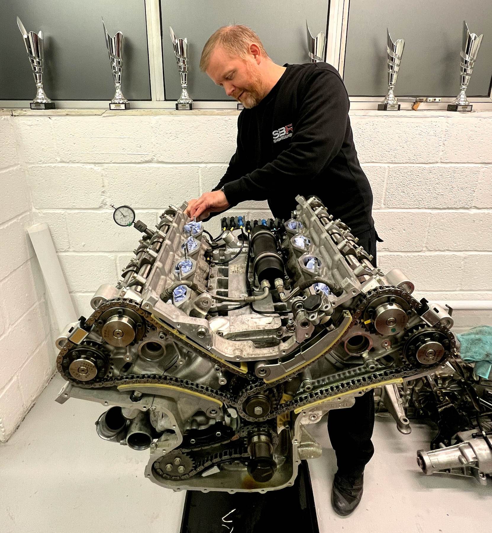 An SBR technician in a black SBR top checking the cam timing on a stripped Ferrari V8 on the engine bench, with race trophies on the shelf behind