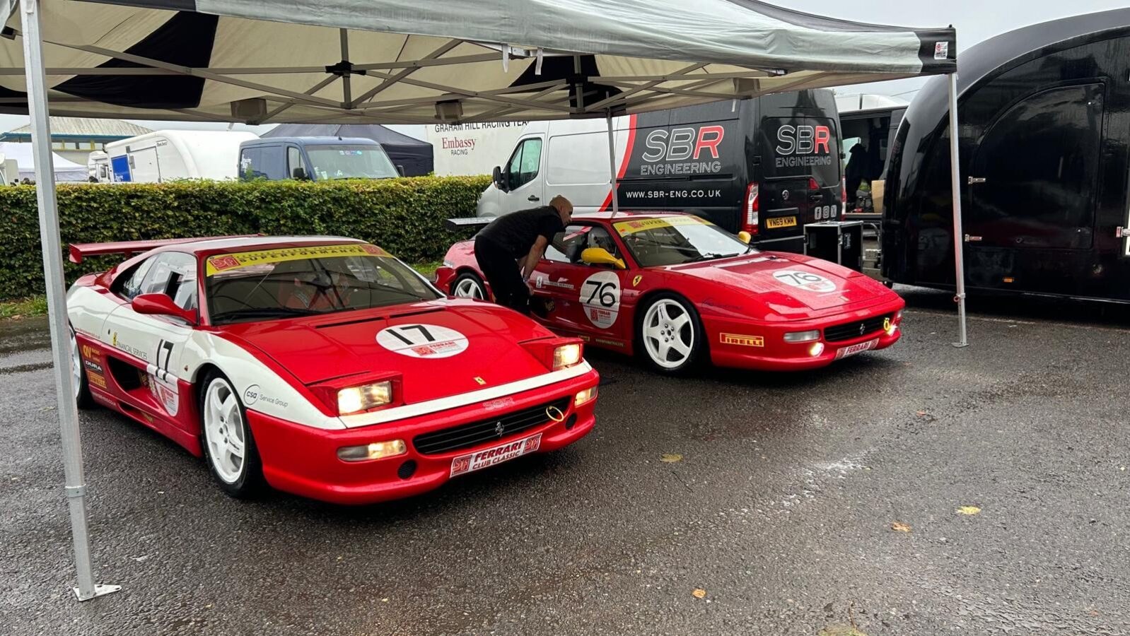 Two red Ferrari F355 Challenge cars, numbers 17 and 76, under the SBR Engineering gazebo in the paddock with the SBR van behind