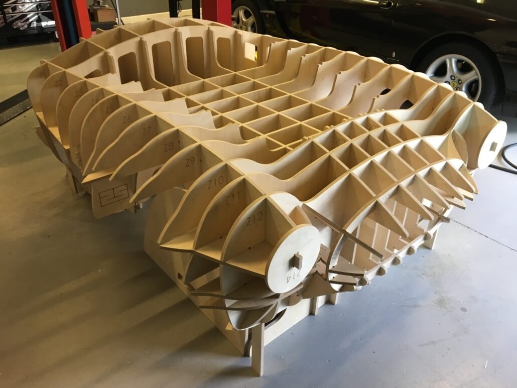 A CNC cut plywood body buck for a Cobra sitting on the workshop floor, with a black Ferrari behind