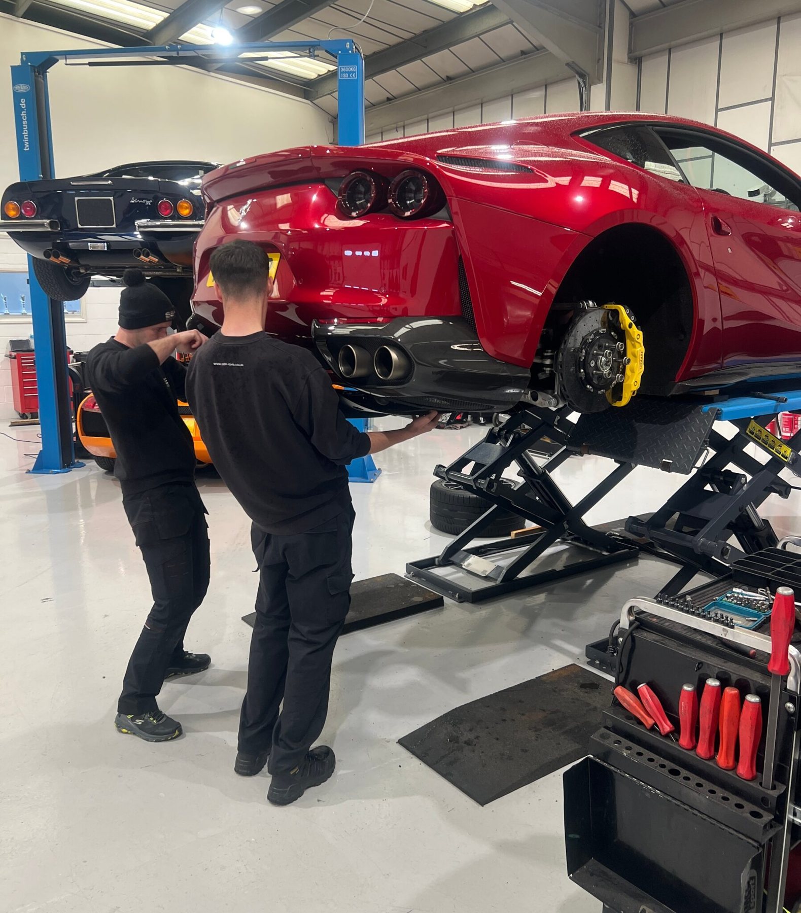 An SBR technician working under a red Ferrari 812 raised on a two post lift in the SBR workshop