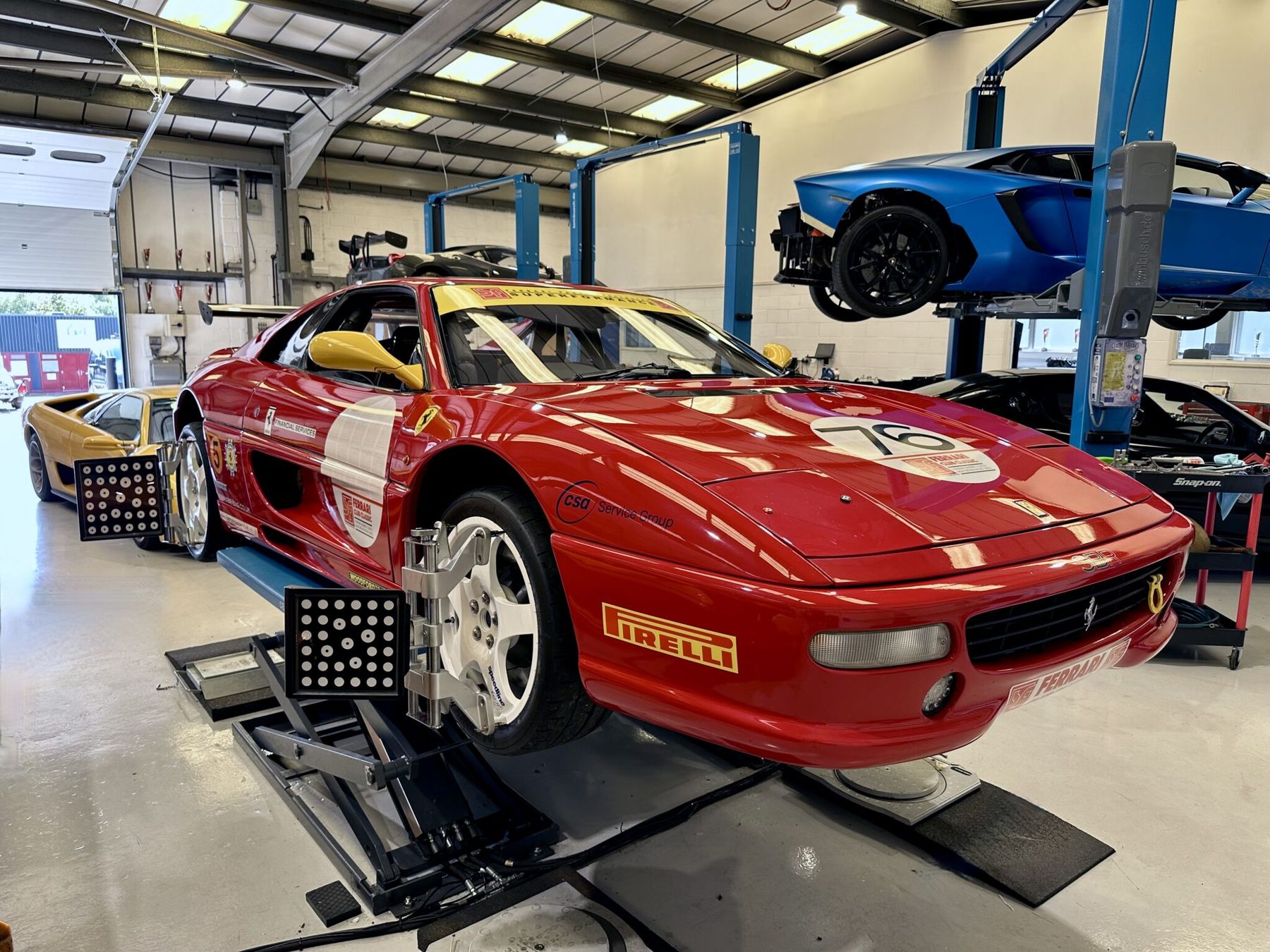 A red Ferrari F355 Challenge, number 76, on the geometry rig with alignment targets fitted to its wheels, and a blue Lamborghini Aventador on the lift behind