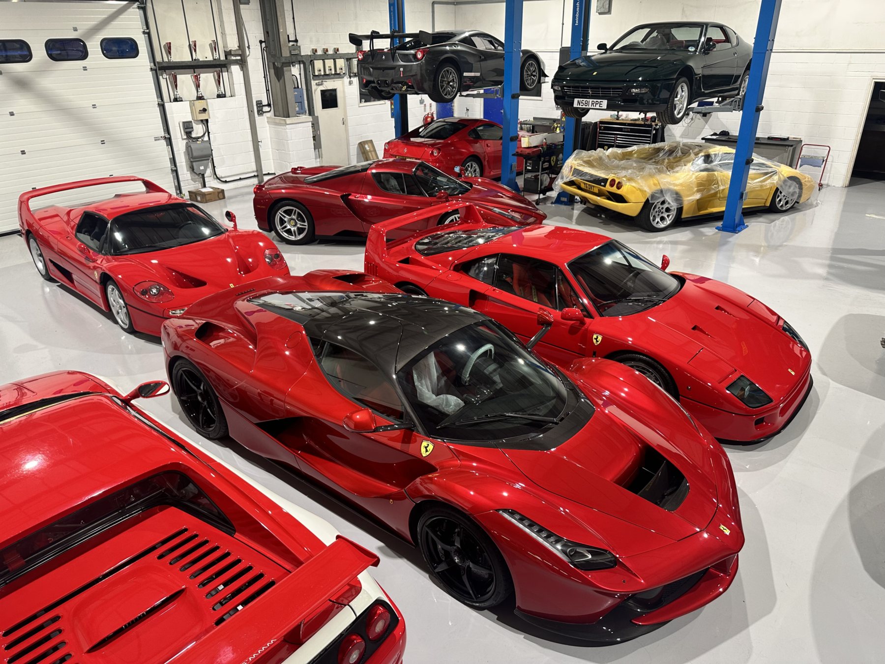 A red Ferrari LaFerrari, F40, F50 and 599 GTO parked together on the white floor of the SBR Engineering workshop, with a 458 Challenge and a Daytona on the lifts behind