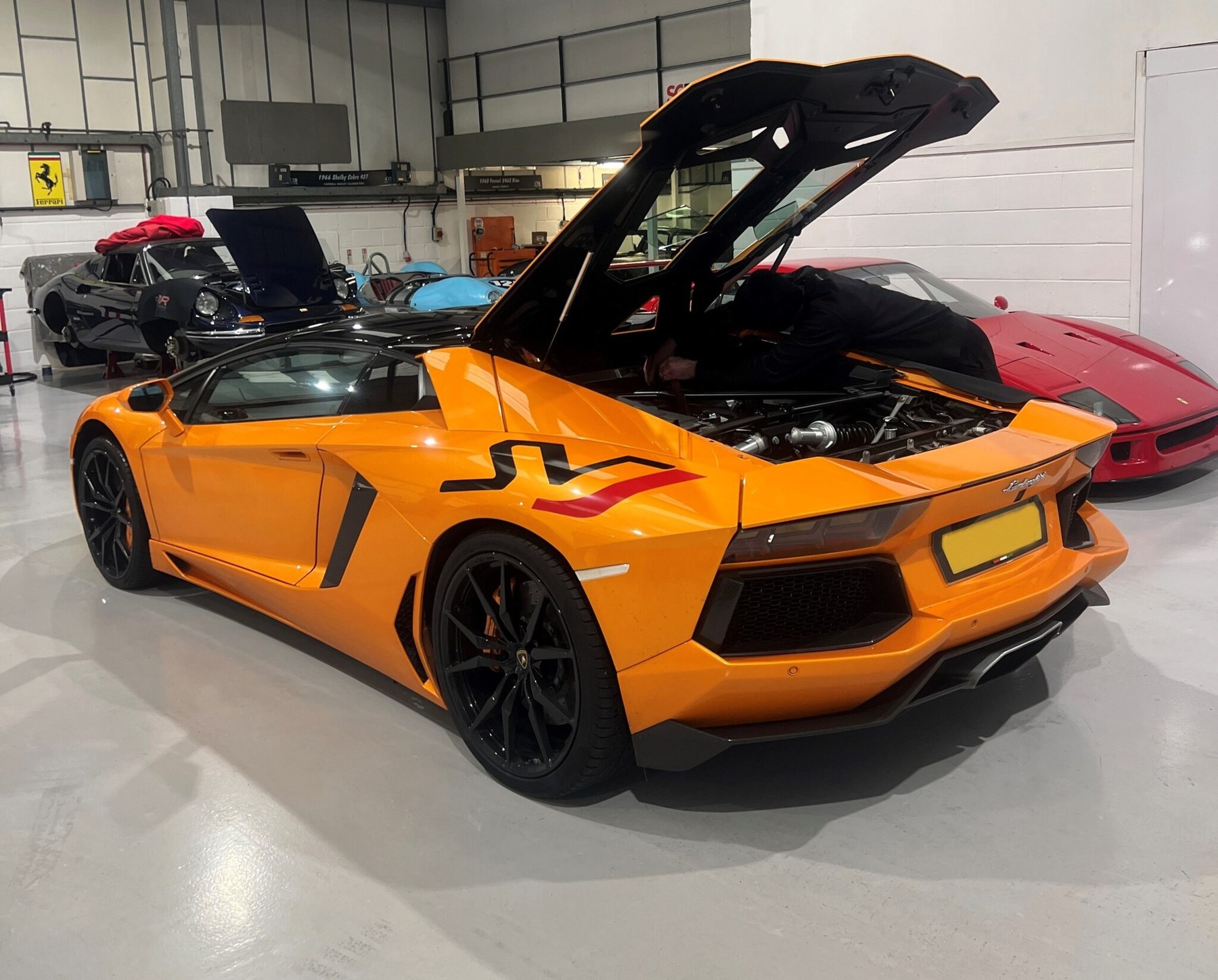 An orange Lamborghini Aventador with its engine cover raised in the SBR workshop, with a red Ferrari F40 and other cars behind