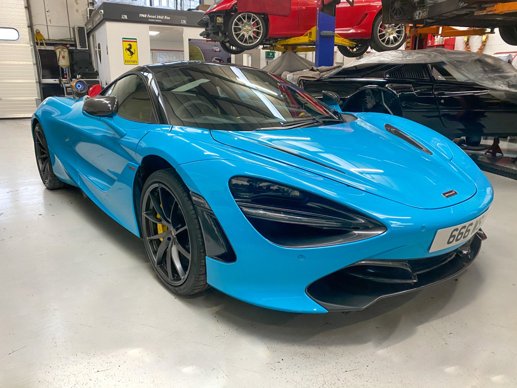 A light blue McLaren 720S on the SBR workshop floor with cars on the lifts behind it