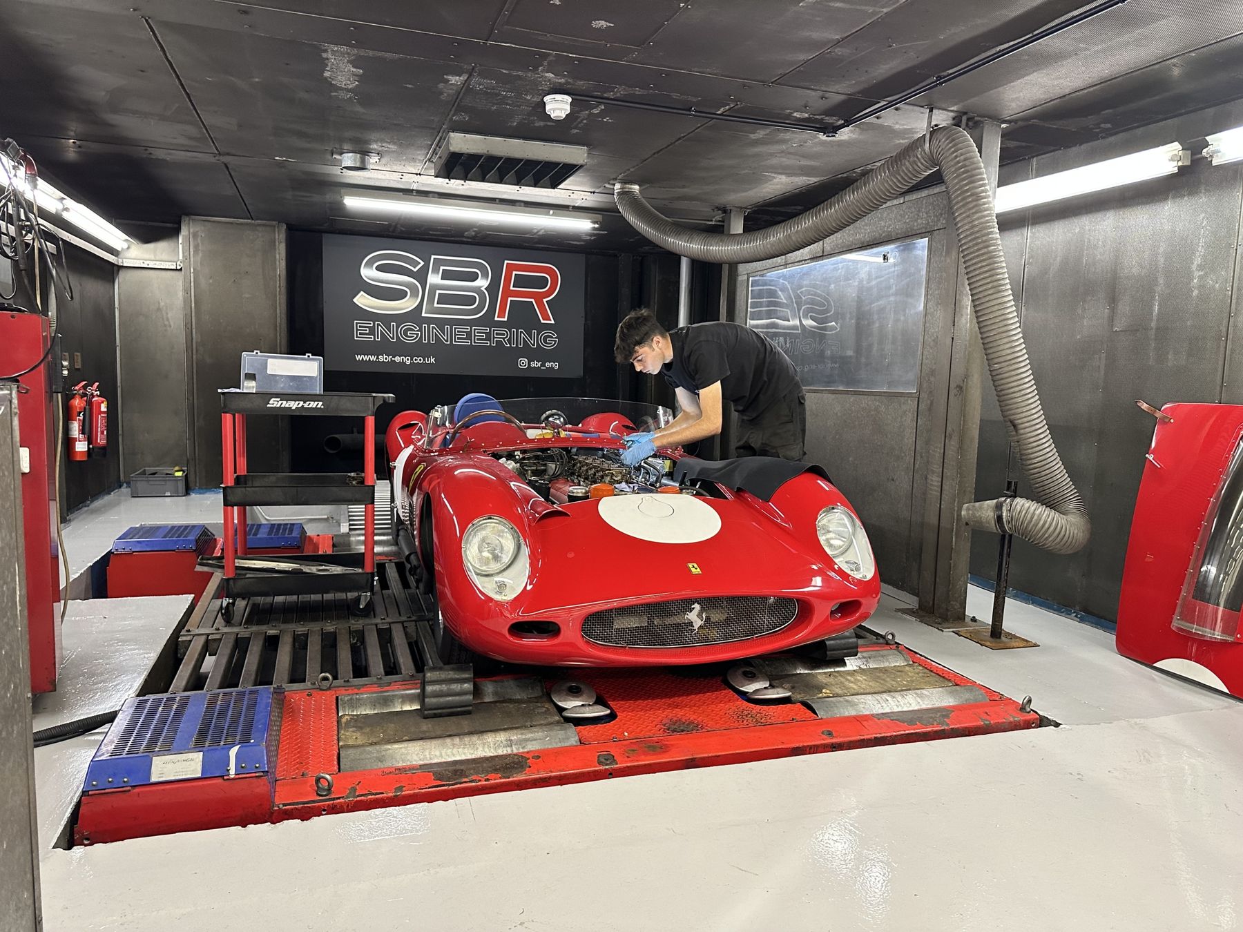 A red Ferrari 250 Testa Rossa style sports racer strapped to the SBR Engineering rolling road, with a technician at the controls and the SBR banner above