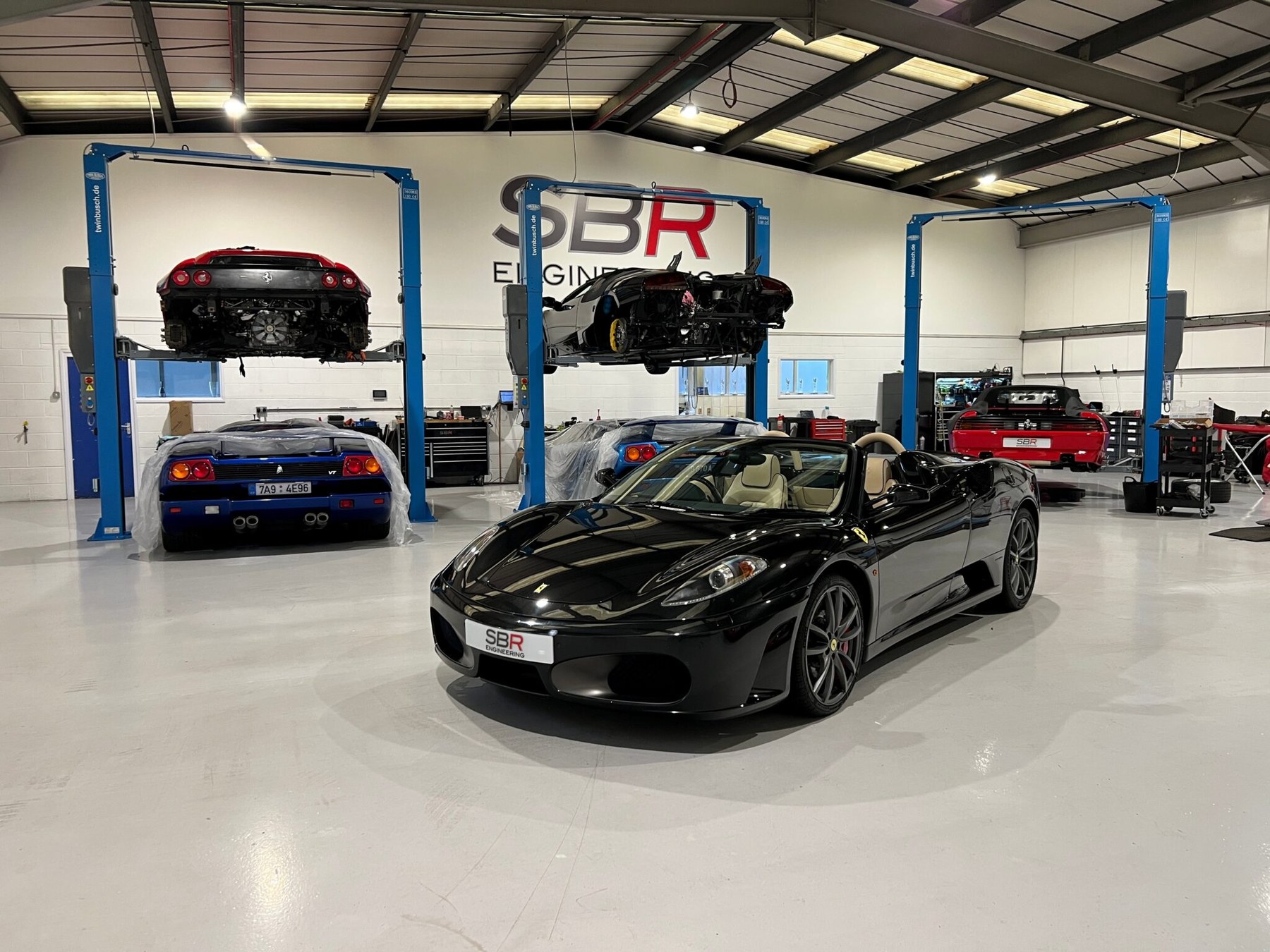 A black Ferrari F430 Spider on the workshop floor at SBR Engineering, with two Ferraris on the lifts and the SBR Engineering sign on the wall behind