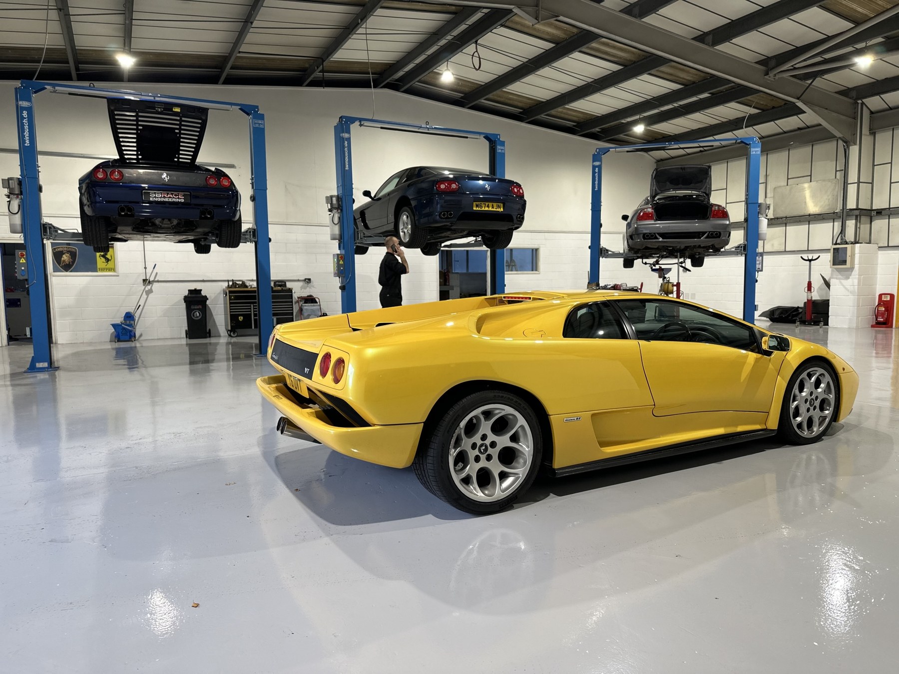 A yellow Lamborghini Diablo 6.0 on the SBR workshop floor with three cars raised on two post lifts behind it